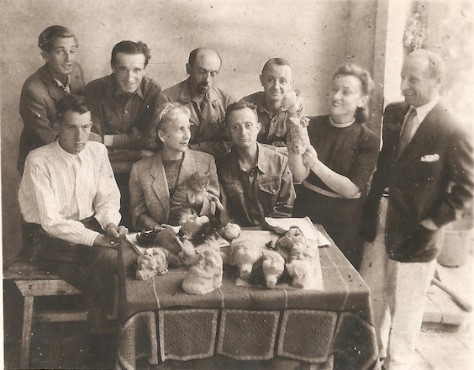 Dziewięć osób, w tym dwie kobiety, zgromadzonych przy stole, na którym leżą odlewy główek lalek. Pośrodku siedzą: młody mężczyzna w białej koszuli, kobieta w średnim wieku, z kotem na kolanach, obok niej drobny mężczyzna w koszuli od munduru. Inni sto