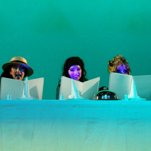 W seledynowym świetle, zza długiego stołu przykrytego obrusem w tym samym kolorze, widać  głowy sześciu kobiet, skupionych na lekturze trzymanych przed sobą białych kartek. 