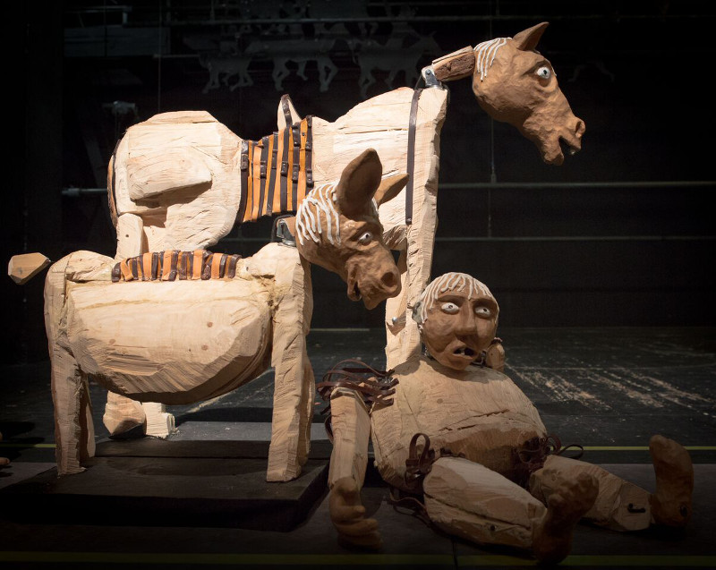 Drewniane, surowe rzeźby lalek: grubasa, osła i konia ustawione na czarnym tle. 