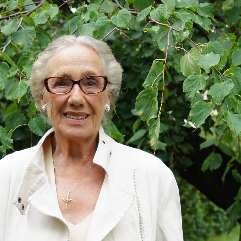 Kolorowy portret siwej, starszej, uśmiechniętej kobiety w okularach, w białej bluzce, na tle zielonych liści. 