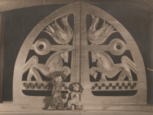 Centralnie, na tle ozdobnej, grubej, otwieranej kraty z motywami koni i ptaków, stoją dwie małe lalki z bujnymi włosami. Zdjęcie w kolorze sepii. 