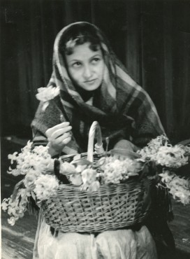 Czarno-biały portret młodej kobiety otulonej grubą kraciastą chustą, spod której wysuwają się na czoło ciemne włosy. Oczy ma skierowane ku górze. W prawej ręce trzyma biały kwiat. Drugą podtrzymuje wiklinowy kosz z kwiatami postawiony na kolanach. 