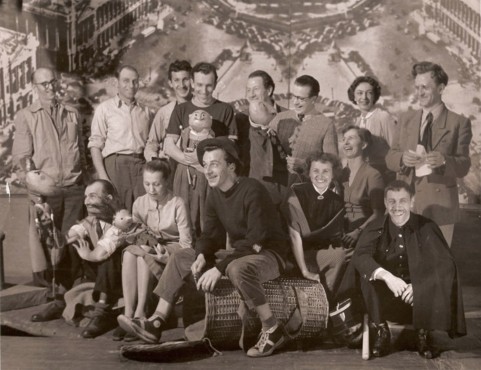 13-osobowy zespół z lalkami na tle powiększonej fotografii Paryża. W pierwszym rzędzie pośrodku, na wiklinowym koszu, młody mężczyzna w czarnym kapeluszu i swetrze. Z jego prawej strony młoda kobieta z lalkę chłopca z czarną czupryną. Sepia. 