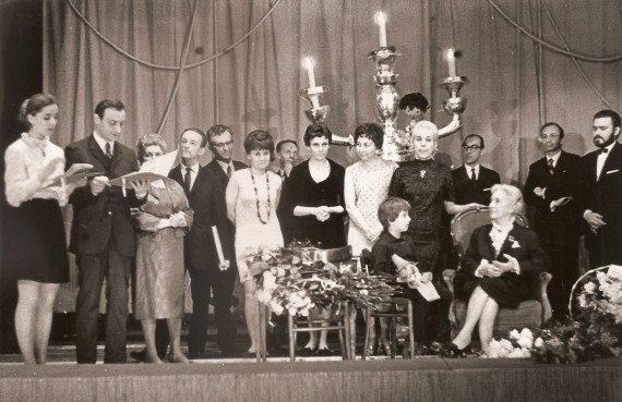 Czarno-białe zdjęcie grupowe na tle kurtyny. Na pierwszym planie: stolik z bukietami kwiatów, obok kilkuletni chłopiec z pacynką skrzata. Z prawej, w fotelu, siwa kobieta w starszym wieku, w jasnej bluzce z żabotem i ciemnym kostiumie. 