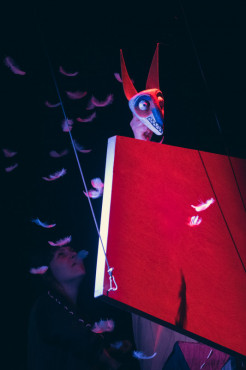 Zza pionowo ustawionej czerwonej platformy wychyla się, wśród fruwających piór, niebiesko-czerwona głowa-maska zwierzęcia o długim pysku, dużych oczach i spiczastych uszach. 