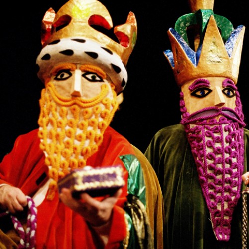 Trzy postacie, jedna za drugą, stoją ubrane w kolorowe maski i brody ze sznurków, w koronach na głowach. 