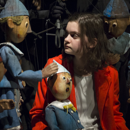 Dziewczynka o ciemnych włosach, w czerwonym płaszczyku, siedzi w otoczeniu trzech pajacyków-marionetek ubranych w niebieskie kostiumy.  