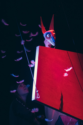 Zza czerwonej platformy zawieszonej na sznurach wychyla się głowa Kuny z czerwonymi uszami i błękitnymi oczami. 