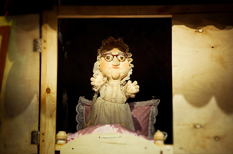W zbliżeniowym ujęciu, z domku wygląda lalkowa postać Babci w okularach, oparta o różową poduszkę.   