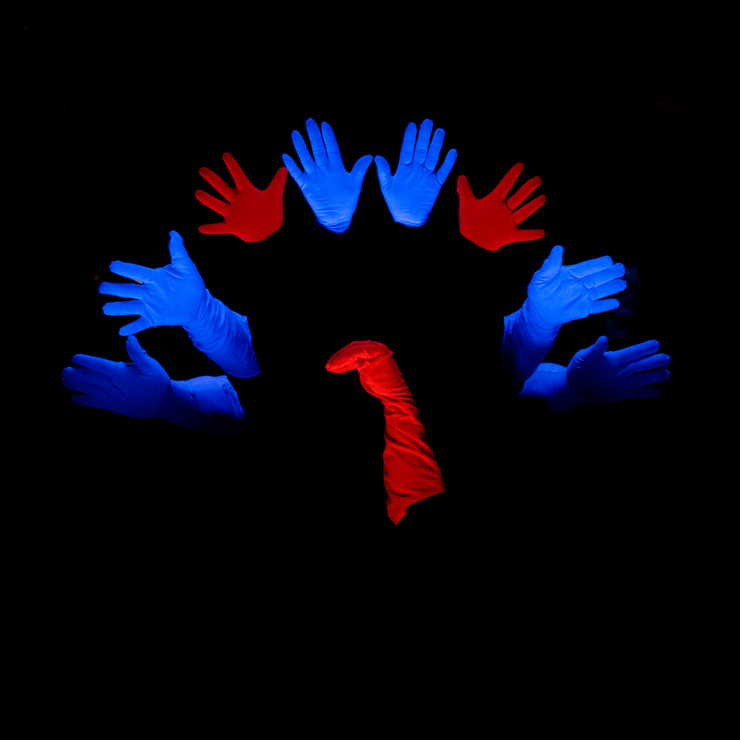 Na czarnym tle ułożony jest wachlarz z niebiesko-czerwonych dłoni. Po środku, widoczna do łokcia, ze zgiętymi palcami dłoni, ręka w czerwonej rękawiczce. 