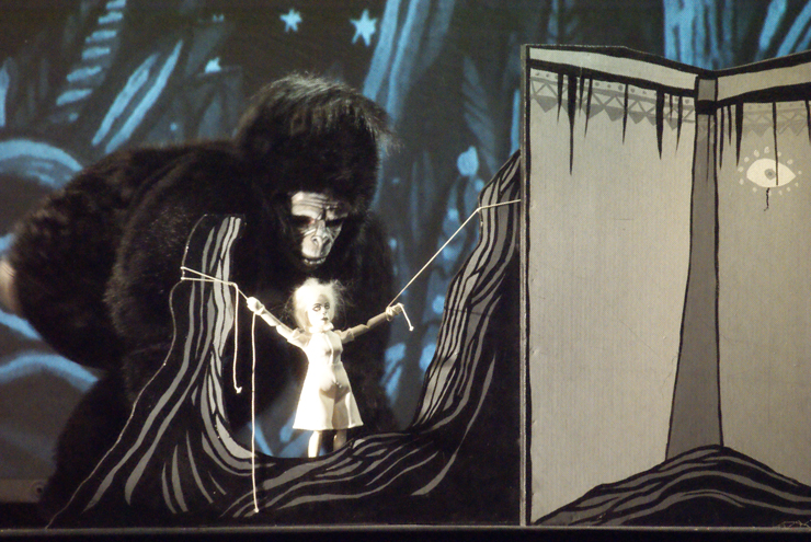 Po lewej stronie wielka małpa (King-Kong) przygląda się wiszącej przed nią małej białej lalce kobiety, przywiązanej za ręce do płaskiej szarej dekoracji. Po prawej szary parawan. W głębi błękitne tło.  