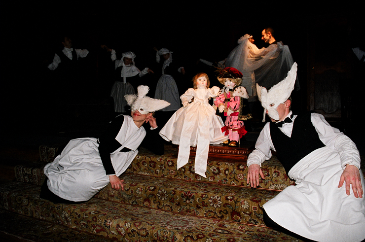Na schodach, po dwóch stronach ustawionej centralnie ślubnej pary dziecięcych lalek, siedzą postacie w białych fartuchach służących i maskach białych gołębi. W tle para młodych tańczy w żywym planie. 