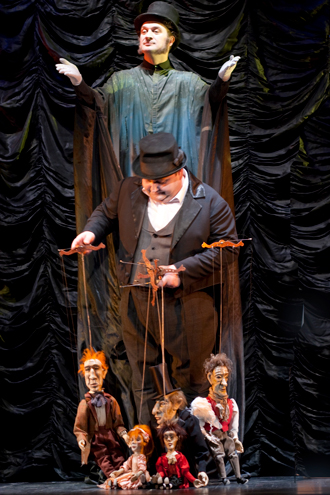 W centralnej części stoi, ubrany na czarno, mężczyzna w cylindrze, który trzyma przed sobą pięć kolorowych marionetek poruszanych na przywiązanych do krzyżaków nitkach. Za nim, na podwyższeniu, na tle udrapowanej czarnej materii, stoi czarna postać.