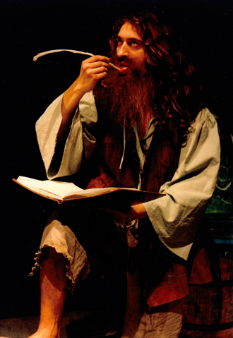 Mężczyzna z długimi włosami i brodą, ubrany w płócienną koszulę i brązowa kamizelkę, siedzi nad otwartą księgą. W ręku trzyma pióro, które zbliża do ust. 