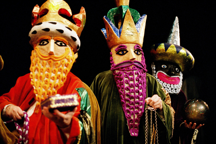 Trzy postacie, jedna za drugą, stoją ubrane w kolorowe maski i brody ze sznurków, w koronach na głowach. 