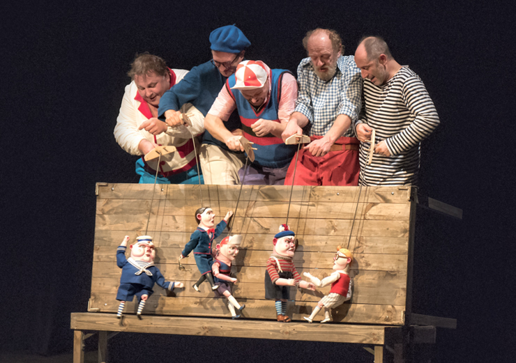 Grupa pięciu mężczyzn, ubranych w marynarskie stroje, w letnich czapkach, animuje na drewnianym podeście marionetki, które odpowiadają wyglądem ludzkim postaciom. 