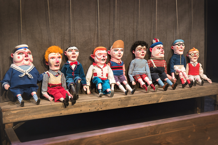 Na drewnianym podeście siedzi dziewięć kolorowych, groteskowych w wyrazie, marionetek chłopców w krótkich spodniach. Niektórzy noszą w marynarskie ubrania, czapki z daszkiem. Inni mają odkryte głowy i bujne czupryny.  