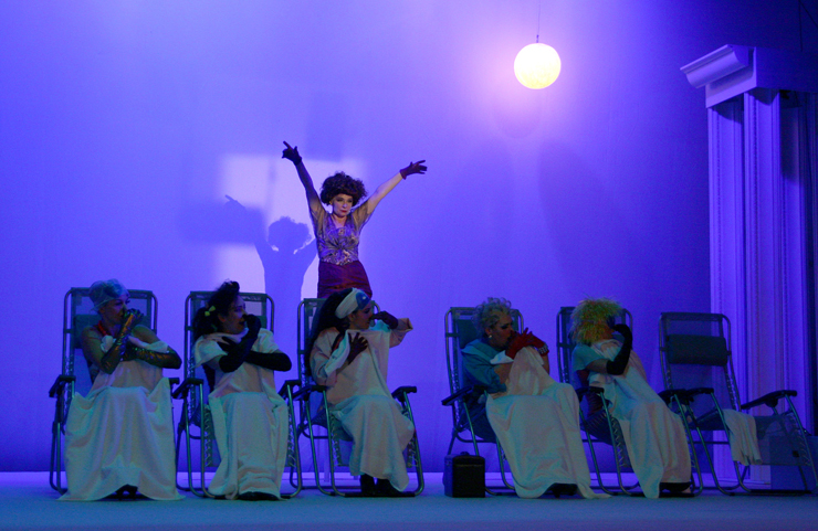 W fioletowym świetle sześć kobiet siedzi na ustawionych w szeregu leżakach. Za nimi, w głębi, znajduje się stojąca w tanecznej pozie postać kobiety w średnim wieku, w plażowej sukience.