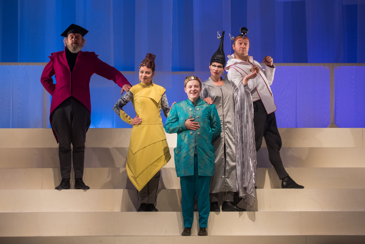 Na błękitnym tle, na kilku stopniach beżowych schodów, stoi pięć osób, od lewej: mężczyzna w czerwonym surducie i czarnych spodniach, kobieta w żółtej sukni , młody mężczyzna w zielonym kostiumie w koronie na głowie, kobieta w srebrnej długiej sukni i wyso