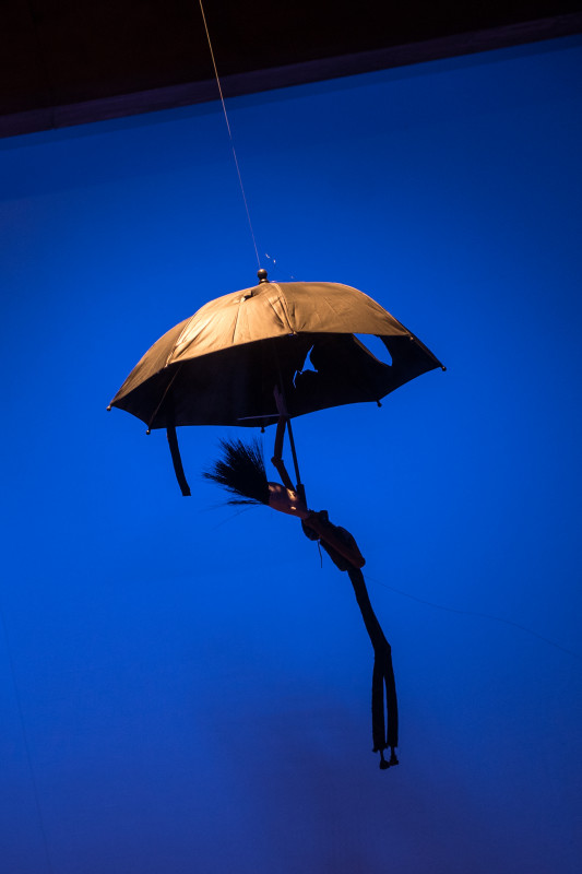 Na błękitnym tle sylwetka unoszącego się w powietrzu parasola z przyczepioną do niego lalką z długimi nogami.   