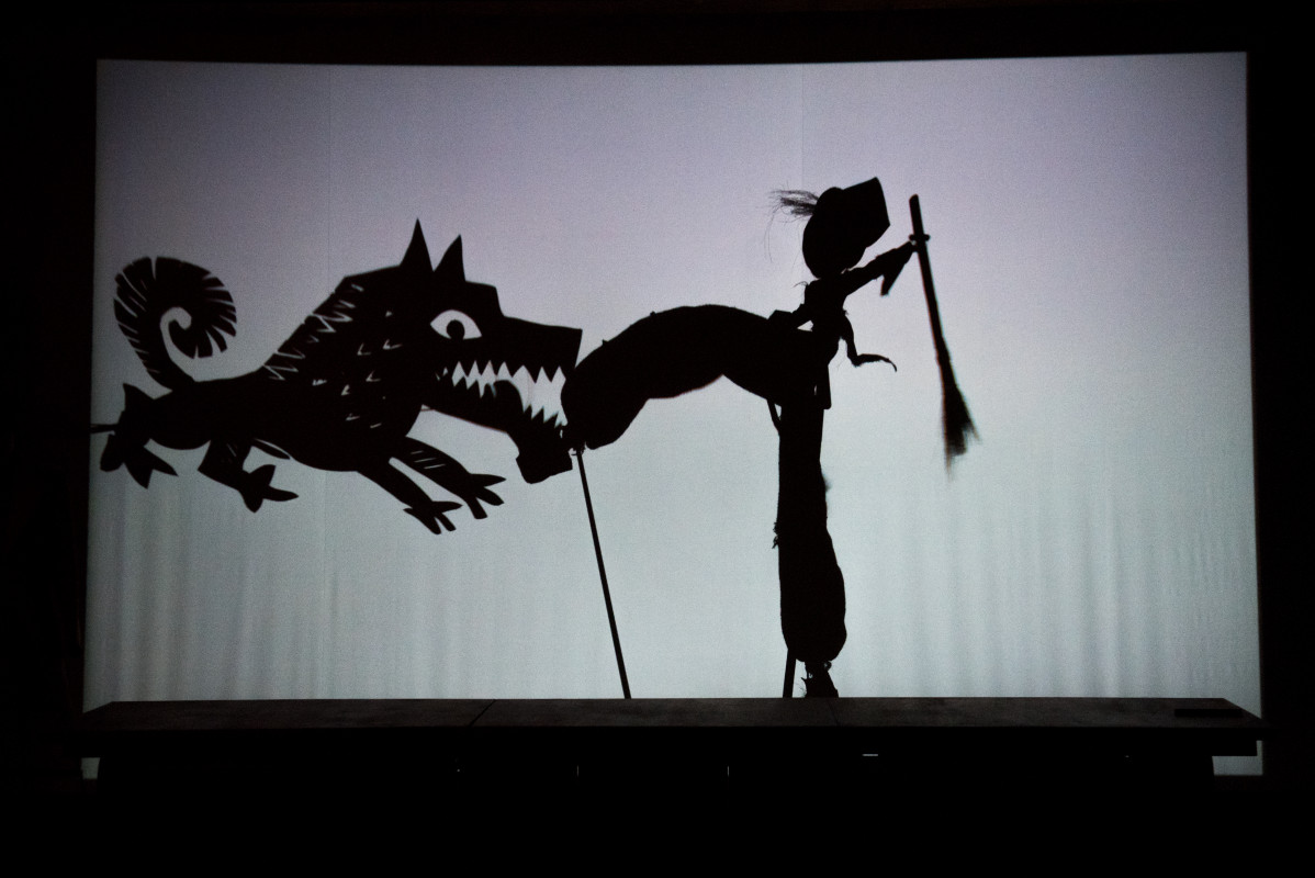 Na białym ekranie czarne cienie: wilka łapiącego za nogę postać w szerokich spodniach i cylindrze. 