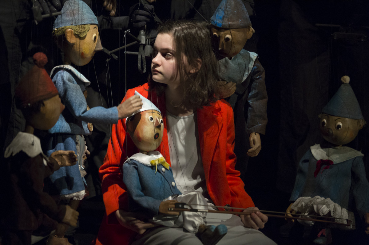 Dziewczynka o ciemnych włosach, w czerwonym płaszczyku, siedzi w otoczeniu trzech pajacyków-marionetek, ubranych w niebieskie kostiumy.  