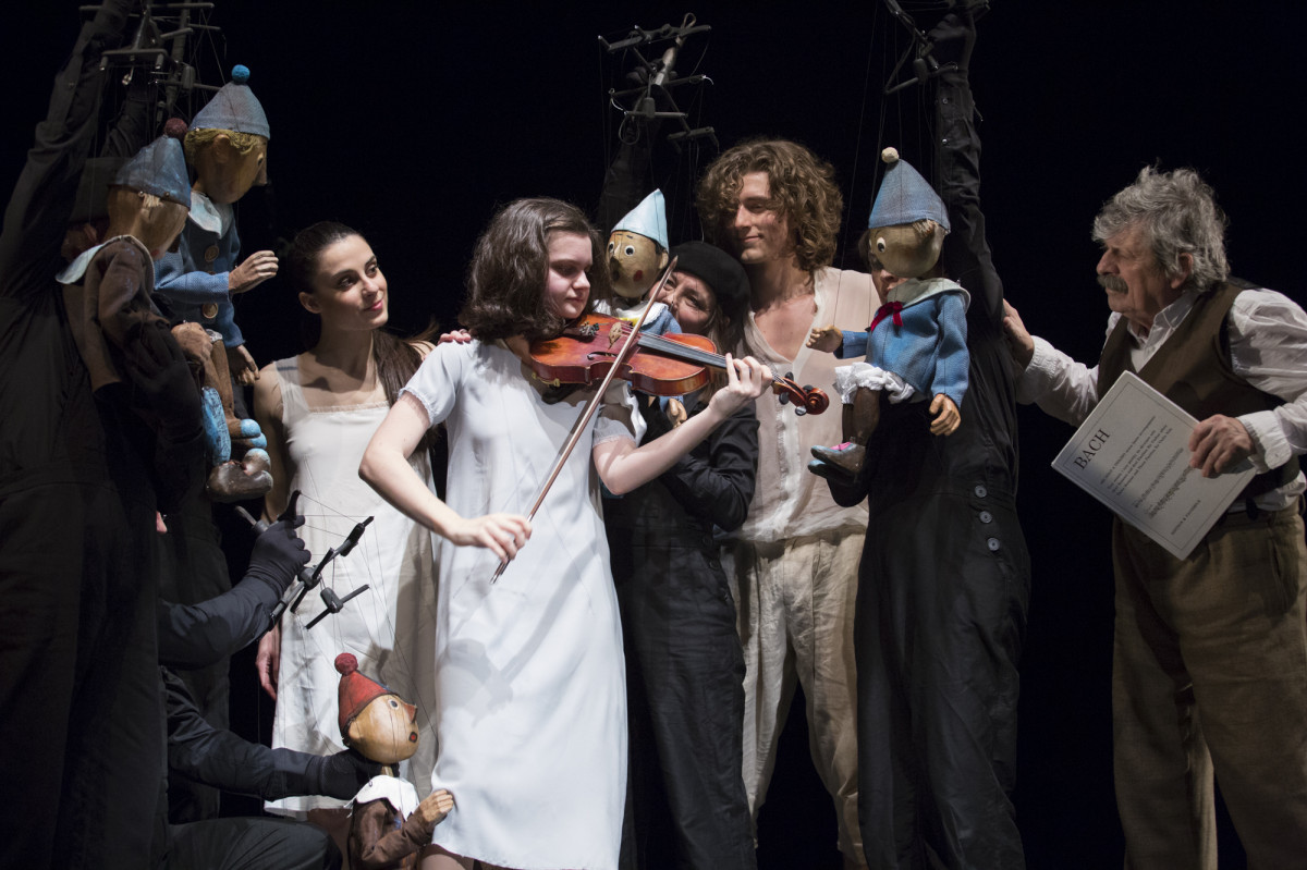 Grupa postaci: po środku dziewczynka w białej sukience grająca na skrzypcach, obok z prawej młody mężczyzna z 2 marionetkami-pajacykami ubranymi na niebiesko, dalej starszy mężczyzna. Z drugiej strony młoda kobieta w białej sukience i jeszcze 3 marionetki.