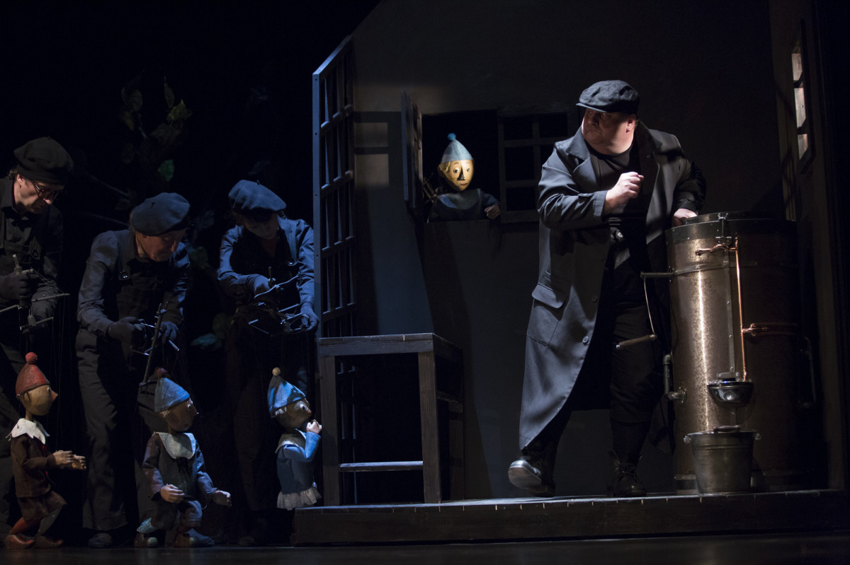 W ciemności widać niebieskie postaci trzech wędrujących pajacyków-marionetek animowanych przez aktorów. Po prawej, obok miedzianego kotła stoi potężny mężczyzna w czarnym płaszczu i czapce. 