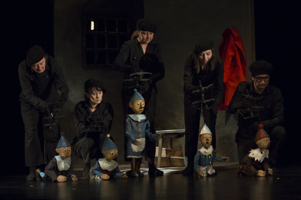 W szeregu stoi lub siedzi 5 pajacyków-marionetek. 4 w niebieskich kostiumach, jedna w brązowym, z czerwoną czapeczką. Podtrzymywane są na nitkach przez ubranych na czarno animatorów.  