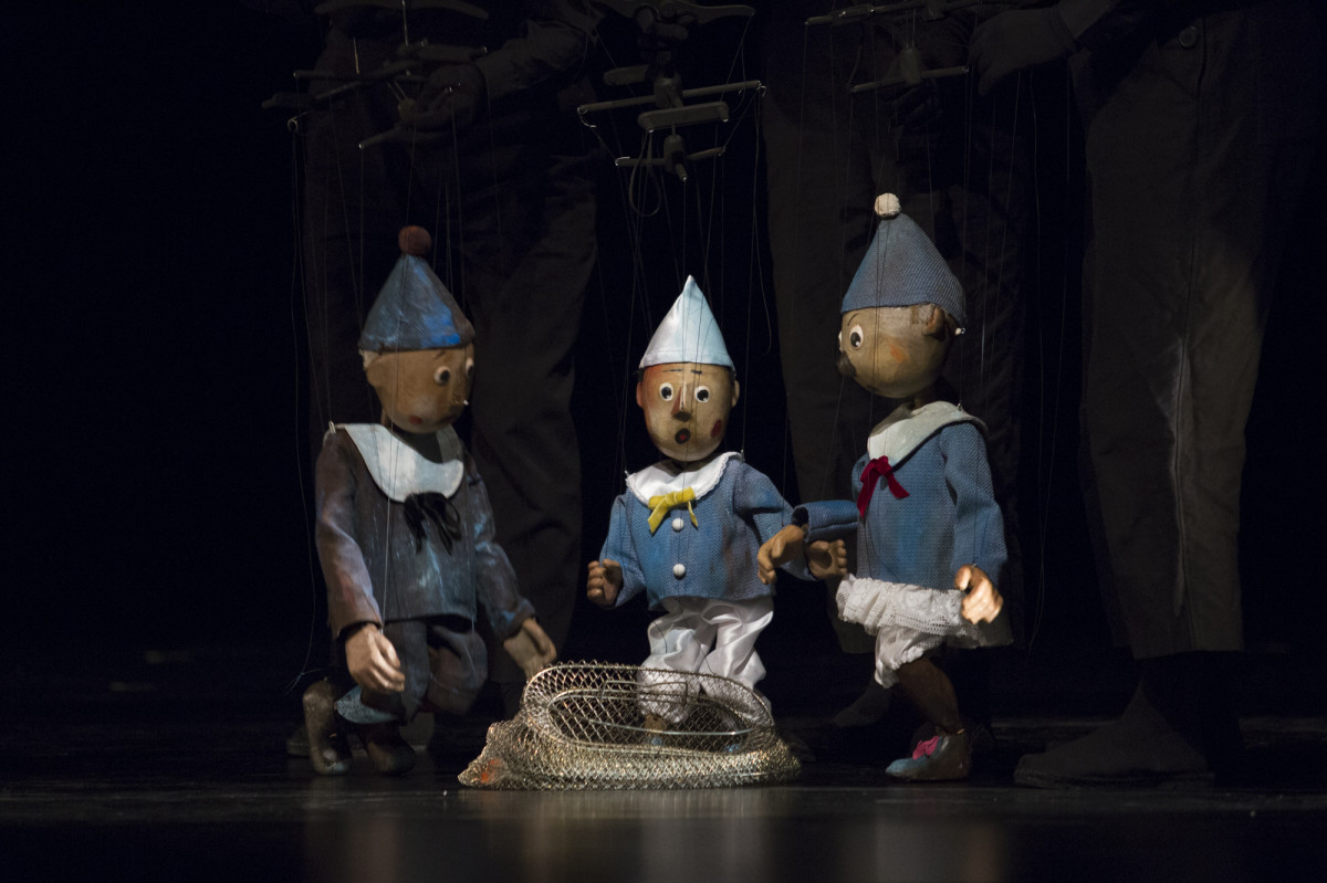 Trzy pajacyki-marionetki w niebieskich kostiumach i czapeczkach, z białymi kołnierzami, stoją nad leżącą na ziemi złotą siecią rybacką. 