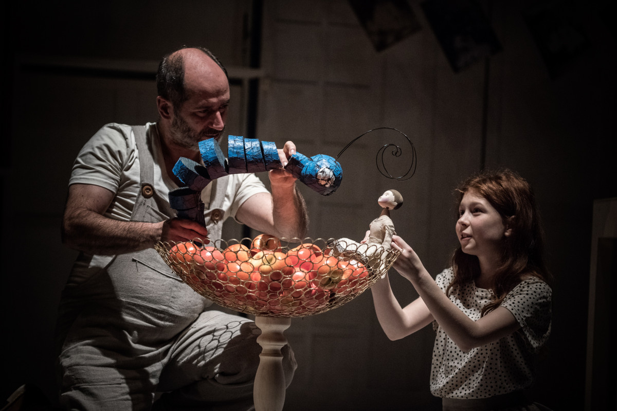 Z prawej strony, nad misą wypełnioną jabłkami, dziewczynka trzyma małą lalkę;  z lewej strony mężczyzna trzyma lalkę błękitnej gąsienicy.